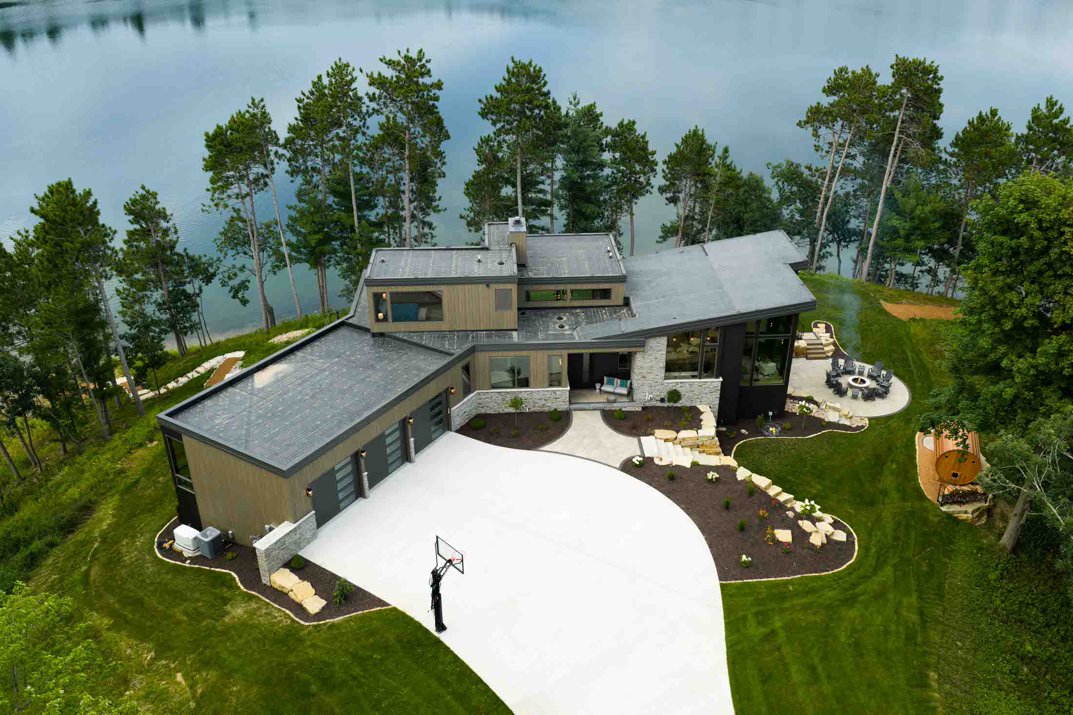 Modern lakeside home featuring a basketball court, landscaped yard, and scenic views of surrounding trees and water.